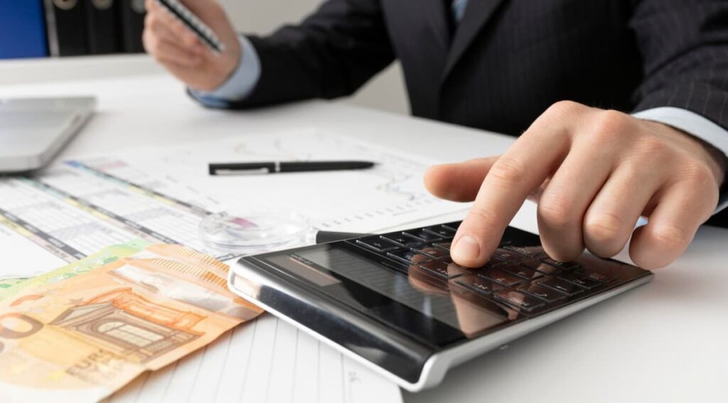 A businessperson uses a calculator on a desk with financial charts and cash