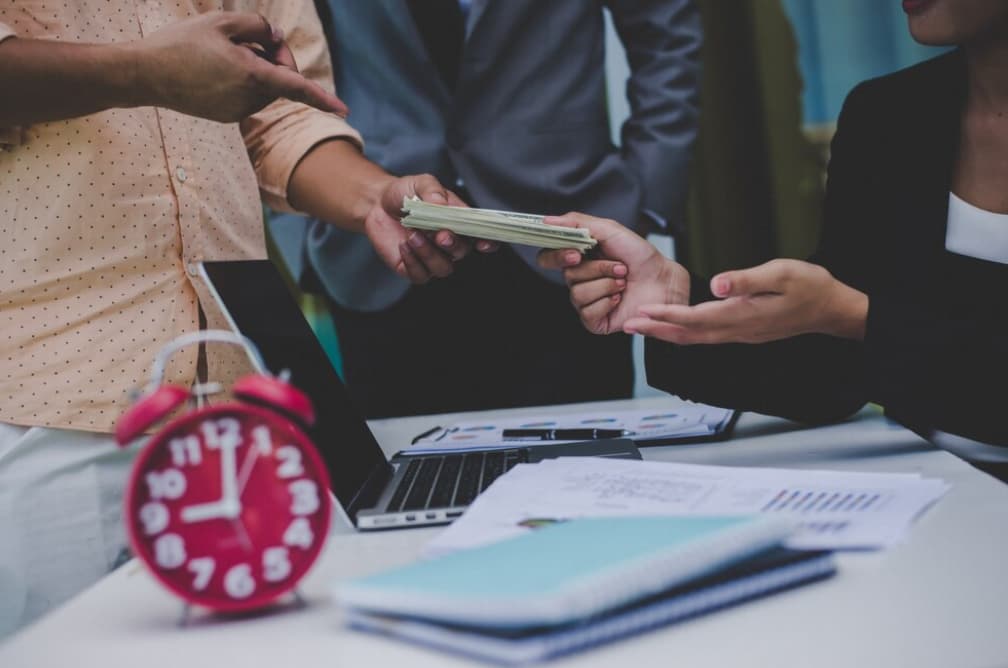 Hands exchanging cash with a clock and laptop on the table