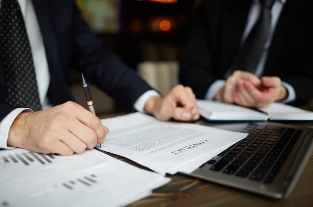 Close-up of hands signing legal documents with a laptop nearby