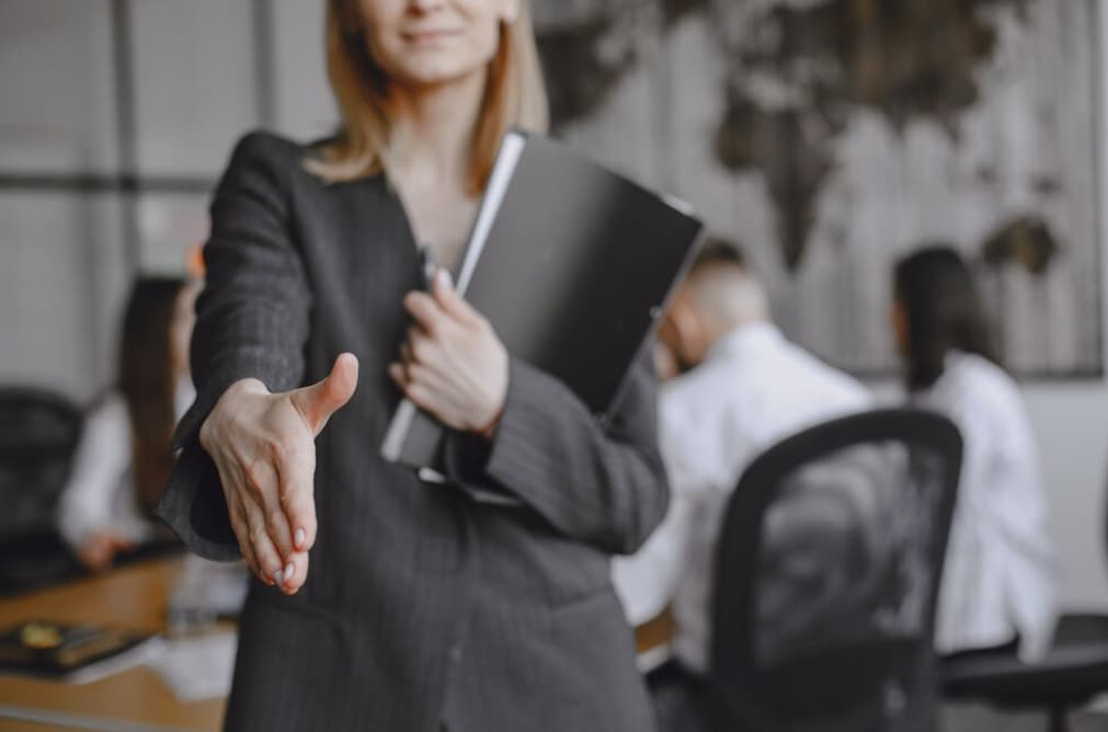 A woman extends a handshake, holding a folder, in an office